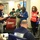 The image depicts an emergency operations center or command post with multiple personnel gathered for what appears to be a briefing or coordination meeting. Staff members are wearing color-coded vests (red, blue, green) indicating different roles or agencies, and several are taking notes. A seated individual in the foreground has a placard reading 'NRCS DEPUTY,' suggesting this is a USDA Natural Resources Conservation Service emergency response operation.