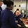 Several U.S. Coast Guard personnel in navy blue uniforms are gathered at a workstation in what appears to be an operations or command center. A seated officer is engaged in conversation with two standing colleagues, one of whom has their back to the camera. Multiple computer monitors, phones, and office equipment are visible in the background, with glass-partitioned rooms bearing partial lettering.