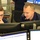 Two uniformed personnel, likely U.S. Coast Guard officers based on their dark blue uniforms and visible rank insignia, are seated at a workstation reviewing information on computer monitors. The individual on the right appears to be speaking or explaining something to the colleague on the left, who is listening attentively with hand raised to face. The setting appears to be an operations or command center with multiple monitors and display screens visible.