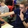A U.S. Coast Guard officer named Bodner, wearing blue duty uniform with captain's bars insignia, works at a computer workstation alongside a civilian woman in a striped sweater who is pointing at the screen. A third individual in Coast Guard uniform is partially visible on the right side of the frame. The setting appears to be an operational command or coordination center.