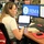 A woman is working at a FEMA operations center, seated at a multi-monitor workstation displaying the FEMA logo and Department of Homeland Security seal. She is using a smartphone and laptop simultaneously while wearing an earpiece, suggesting active coordination during an emergency management operation. A red vest with partial text visible is draped over her chair, and a person in military uniform is visible in the background.