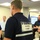 The image shows an emergency operations center or command post with multiple personnel present. The central figure, seen from behind, wears a navy vest with a FEMA-issued identification label reading 'NRCS CHIEF.' Personnel in various roles, including someone in a red safety vest, are gathered around workstations with monitors and a digital display board visible.