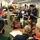 The image shows a busy emergency operations center (EOC) with multiple personnel gathered for a briefing or coordination meeting. Staff members are wearing various identifying vests and badges, including Red Cross volunteers, military personnel in camouflage uniforms, and civilian emergency management officials. The setting appears to be an active emergency response operation with workstations, computers, and documents visible throughout.