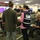 The image shows a group of professionals gathered in what appears to be an emergency operations center or command center. Multiple individuals are engaged in discussions, with one person visibly wearing a 'Tribal Affairs Advisor' vest, suggesting this is an emergency management exercise or real incident response. The room features digital display boards, computer workstations, and red LED clocks showing time zone indicators.
