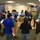A group of approximately 20 people are gathered in what appears to be an emergency operations center or command room, standing in a loose circular formation. Several participants are wearing blue safety/identification vests, suggesting an official drill, exercise, or briefing session. The setting includes institutional lighting, a digital display board, and signage indicating a secured facility.