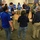 A group of approximately 15-20 people are gathered in a circle in what appears to be an office or training room. Several individuals are wearing blue safety/emergency management vests, suggesting this is a briefing, training exercise, or emergency preparedness meeting. Some participants are holding notebooks or papers, indicating note-taking or reference materials are in use.