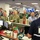 A briefing or coordination meeting is taking place in what appears to be an emergency operations center, likely a FEMA facility. Personnel including military officers in Army combat uniforms and civilian staff wearing ID badges are gathered around a workstation. The individual in the foreground wears a vest labeled 'NRCS CHIEF' with a FEMA logo.