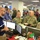 The image shows a busy emergency operations center (EOC) with civilian and military personnel gathered around workstations. Two U.S. Army soldiers in camouflage uniforms are consulting with civilian staff, likely during an emergency management briefing or coordination meeting. Multiple computer monitors, display screens showing maps and data, and a crowded workspace suggest an active emergency response operation.