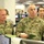 The image shows U.S. Army personnel in an operations or command center environment, wearing OCP (Operational Camouflage Pattern) uniforms. Two primary soldiers are visible in the foreground — one identified by name tape as 'YOUNG' holding a notebook, and another partially visible with name tape 'ROSE.' A female Air Force member is visible in the background along with several civilians.