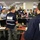 A briefing or coordination meeting is taking place in what appears to be an emergency operations center (EOC). Personnel from multiple agencies, including FEMA and NRCS, are gathered around workstations listening to a speaker. The setting includes multiple monitors, display screens showing maps and weather data, and a large group of mixed civilian and military personnel.