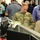 A busy operations or command center environment where military personnel and civilians are gathered around workstations. A U.S. Army soldier with the name tape 'ROSE' in camouflage uniform is engaged in conversation with a civilian male and a woman in a tan blazer. Multiple computer monitors and workstations are visible, suggesting a coordination or briefing scenario.