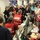 The image depicts a busy emergency operations center (EOC) with a large group of personnel gathered for what appears to be a briefing or coordination meeting. Several individuals are wearing red vests with labeled roles such as 'Resources &amp; Capability Branch Director' and 'Resource Support Section Deputy,' indicating a structured incident command system. The setting includes multiple workstations with computers, monitors, and communication equipment consistent with a FEMA or similar emergency management facility.