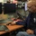 A FEMA employee is working at a multi-monitor workstation in what appears to be an emergency operations center. The man, wearing a navy blue FEMA jacket and glasses, is using a laptop while a desktop computer with dual monitors is also present at his station. The background shows additional personnel and workstations consistent with an active operations center environment.