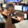 The image shows a FEMA operations center or emergency management facility where multiple personnel are working at computer workstations. Two individuals in the foreground appear to be engaged in a conversation, with one gesturing expressively toward the other. The room contains numerous monitors displaying FEMA branding and operational data, suggesting an active emergency coordination environment.