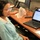 A woman wearing glasses and a blue-and-white striped shirt is seated at a wooden office desk working on a Dell laptop. The laptop screen displays what appears to be an email client or document application. The workspace includes multiple monitors, a desk phone, keyboard, mouse, and a plastic shopping bag on the desk.