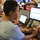 The image shows a busy office or operations center environment with multiple workers at computer workstations. A woman in the foreground wearing a light blue denim top and hoop earrings is actively typing on a keyboard while looking at dual Dell monitors. The background shows additional staff members working at similar multi-monitor setups in what appears to be a command center or call center setting.