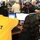 The image shows a busy emergency operations center or command center with multiple personnel working at computer workstations. A person wearing a bright yellow 'IT Support Staff' jacket is assisting another worker wearing a headset at a computer terminal. The room is densely populated with staff, including at least one military personnel in camouflage uniform visible in the background.