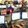 The image shows a busy emergency operations center or command center with numerous personnel working at computer workstations. Multiple rows of desks are equipped with dual monitors, phones, and labeled placards indicating organizational roles. The setting appears to be a FEMA or similar emergency management facility, evidenced by visible signage including 'VAL Unit Leader' and 'ARC LNO' placards.
