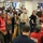 The image shows a busy FEMA (Federal Emergency Management Agency) operations center with numerous personnel gathered for what appears to be a briefing or coordination meeting. Multiple staff members are wearing red FEMA vests with role designations, including 'Resources &amp; Capability Branch Director' and 'Movement Coordination Leader.' The room is filled with computer workstations, monitors displaying data, and whiteboards, indicating an active emergency management operation.