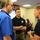 Three men are engaged in a discussion inside what appears to be a FEMA emergency operations center. The room features multiple digital clocks displaying times for different U.S. time zones, computer workstations, and large display screens showing operational data. The central figure is wearing a black FEMA polo shirt, suggesting this is an official emergency management briefing or coordination meeting.
