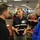 Three men are engaged in a discussion at what appears to be a FEMA emergency operations center. The central figure is wearing a vest labeled 'NRCS Chief' and appears to be briefing or explaining something to the other two individuals. The background shows a busy operations floor with multiple workstations, computer monitors, and personnel.