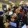 The image shows a busy emergency operations center (EOC) or command center with multiple personnel working at computer workstations. Staff members are engaged in various activities including working on laptops and desktop computers, reviewing documents, and holding discussions. The environment suggests an active emergency management or disaster response operation.