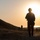 3rd ISOF FID training A silhouetted armed soldier wearing a helmet and carrying equipment stands on a dirt path at sunset or sunrise, overlooking a landscape with a body of water in the background. Several other figures are visible in the distance along the same path. The strong backlight from the sun creates dramatic silhouette effects across the entire scene.