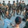 3rd ISOF FID training A group of armed military personnel in desert camouflage uniforms are gathered in a semicircle on sandy terrain, appearing to receive a briefing or instruction from individuals seated or crouching in the foreground. The soldiers are equipped with tactical gear, body armor, and assault rifles. The setting appears to be an outdoor training or operational briefing in an arid environment.