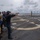 USS MOMSEN Conducts Live Fire Gun Shoot at Sea Military personnel are conducting a live-fire pistol qualification exercise on the flight deck of a naval vessel at sea. Multiple shooters are lined up at a firing line aiming semi-automatic pistols at human silhouette paper targets mounted on stands at the far end of the deck. Observers wearing hearing protection and safety vests stand behind the active shooters.