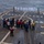 USS MOMSEN Conducts Live Fire Gun Shoot at Sea A group of naval personnel are conducting a pistol qualification or firearms training exercise on the flight deck of a naval vessel at sea. Multiple shooters are standing in a line aiming handguns at white silhouette targets positioned at the far end of the deck. Safety observers wearing colored vests are present to supervise the exercise.