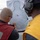 USS MOMSEN Conducts Live Fire Gun Shoot at Sea Two individuals are examining a paper shooting target with multiple bullet holes clustered in the torso area. One person in a red vest is marking or scoring the target with a pen, while another wearing yellow and ear protection observes. The setting appears to be an outdoor firearms training exercise, likely on a naval vessel based on the 'DDG 92' text visible on the cap.