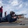 A military or naval personnel member is in a prone shooting position on what appears to be the deck of a naval vessel, aiming an M4/M16-style rifle. Several other personnel in uniform are standing nearby observing, with safety equipment visible. The setting appears to be a shipboard weapons qualification or training exercise conducted at sea.