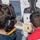 Two individuals are examining shooting targets on what appears to be the deck of a naval vessel at sea. One person is wearing hearing protection (ear muffs branded 'IMPACT') and a yellow-accented uniform, while the other wears a red shirt with the number '16'. Multiple target types are visible, including a standard silhouette target and zeroing targets.