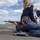 A military or naval personnel member is in a prone shooting position on what appears to be the deck of a naval vessel, aiming a rifle (likely an M16/M4 variant) toward the ocean. Several other personnel are present, some wearing red safety vests and hearing protection, suggesting a supervised live-fire training exercise. Multiple spare magazines are visible on the deck beside the shooter.