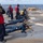 Military personnel are conducting a live-fire rifle qualification exercise on the flight deck of a naval vessel at sea. Multiple sailors are in the prone position firing rifles toward the ocean, while safety observers in red and yellow vests monitor the exercise. Spent brass casings are visible on the deck, and one sailor is using binoculars to observe downrange.