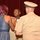 A military promotion or award ceremony is taking place in a formal indoor setting with a red curtain backdrop. A woman in a blue dress is pinning an insignia or device onto a servicemember in a white uniform shirt, while a senior officer in tan/khaki uniform stands nearby. Additional uniformed personnel are visible seated in the background.
