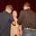 A military promotion or pinning ceremony is taking place in what appears to be an auditorium or formal venue. A service member in tan/khaki military uniform is having insignia or rank devices pinned onto their collar or shoulders by two civilians. An American flag is partially visible in the background alongside a red curtain.