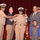 A military promotion ceremony is taking place on a stage with a red curtain backdrop. A Navy service member in the center is having rank insignia pinned to their shoulders by two individuals, while family members including a man in a suit and a young boy stand nearby. The atmosphere appears celebratory, with the honoree smiling broadly.