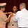 A military promotion or pinning ceremony is taking place in front of a red curtain backdrop. A uniformed service member and a civilian in a white shirt are simultaneously pinning insignia onto the collar or chest of a smiling recipient in a light-colored military uniform. The recipient's joyful expression suggests this is a significant milestone event.