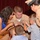 A military promotion or pinning ceremony is taking place, with a male Navy sailor in khaki uniform being pinned by family members and a fellow service member. A woman in a blue dress, two young boys, and a female Navy sailor in uniform are all participating in attaching insignia to the sailor's collar or uniform. The event appears to be held in a formal venue with a red curtain backdrop.