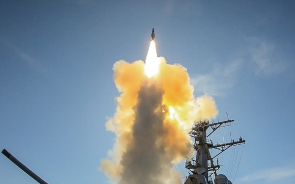 A naval warship, consistent with a U.S. Navy Arleigh Burke-class guided-missile destroyer, is launching a missile from its Vertical Launch System (VLS). A large plume of white smoke and orange exhaust gases billows around the ship as the missile ascends into a clear blue sky. The ship's forward 5-inch deck gun is prominently visible in the foreground.