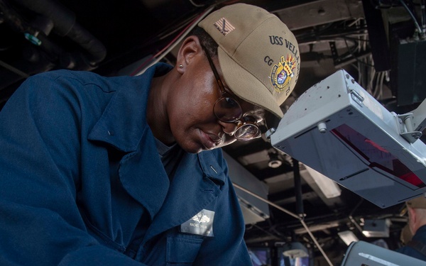 A U.S. Navy sailor wearing a USS Vella Gulf (CG-72) cap and blue coveralls is working at a navigation or chart table aboard a naval vessel. She is using a drafting instrument, likely a compass or divider, on documents or charts. A second crew member is visible in the background working at another station.