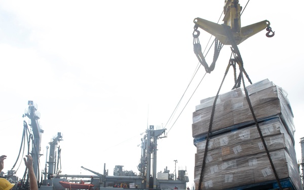 A replenishment-at-sea (RAS) operation is underway between two naval vessels on open, choppy water. A large pallet of boxed supplies is being transferred via crane and rigging from a supply ship to a receiving vessel. Personnel on deck, wearing hard hats and safety gear, are observing and guiding the transfer.