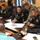 A military planning meeting is taking place around a large conference table, involving personnel in black Thai military uniforms and at least one individual in US Army multicam/OCP camouflage uniform. Participants are reviewing what appears to be a map or architectural floor plan spread on the table. The setting suggests a joint military coordination or briefing session.