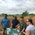 A group of five individuals are gathered outdoors in a field or grassland area, examining what appears to be a map or aerial photograph mounted on a board. The group includes at least one person in military camouflage uniform and one wearing a military-style cap, alongside civilians. They appear to be conducting a field survey, reconnaissance, or site assessment.