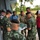 A mixed group of military personnel and civilians are gathered outdoors near a building, reviewing what appears to be a map or aerial photograph. The group includes soldiers in two distinct camouflage uniforms — one pattern consistent with US Army MultiCam and another with a pixelated green pattern — along with several civilians. The meeting appears to be a planning or briefing session in a tropical environment.