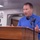 11 MEU, USS Boxer Marines, Sailors pay homage to victims of 9/11 attacks A middle-aged man in a blue long-sleeve shirt is speaking at a wooden podium equipped with a microphone. The setting appears to be aboard a naval vessel, as indicated by a sign reading 'USS BO...' visible in the background. The speaker is gesturing with both hands, suggesting an active and engaged presentation.
