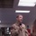 11 MEU, USS Boxer Marines, Sailors pay homage to victims of 9/11 attacks A military personnel member wearing a tan/khaki flight suit with multiple unit patches is speaking into a microphone, likely addressing an audience. The setting appears to be the interior of a military aircraft, possibly a cargo or transport plane. The image is rotated 90 degrees counterclockwise.