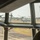 The image is taken from inside a cockpit or flight deck, looking out through multi-pane windows at a military airfield. Multiple MV-22 Osprey tiltrotor aircraft are visible on the tarmac with their rotors spinning. A crew member wearing a flight helmet and headset is visible in the right foreground.