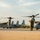 A military airfield scene showing multiple MV-22 Osprey tiltrotor aircraft parked on a tarmac in what appears to be a tropical location. A single military personnel in flight suit stands facing the aircraft in a saluting or shielding-eyes gesture. The warm golden light suggests either early morning or late afternoon conditions.